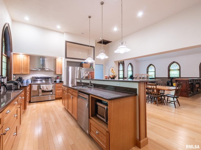 kitchen with light wood finished floors, a towering ceiling, stainless steel appliances, a sink, and wall chimney range hood