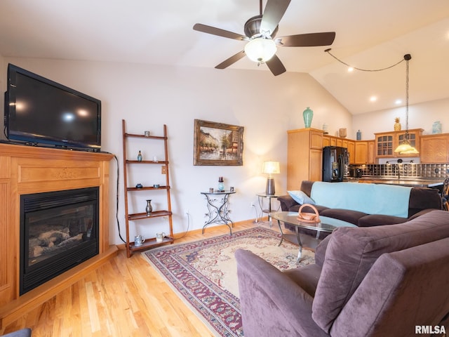 living area featuring a glass covered fireplace, light wood-style flooring, vaulted ceiling, and ceiling fan