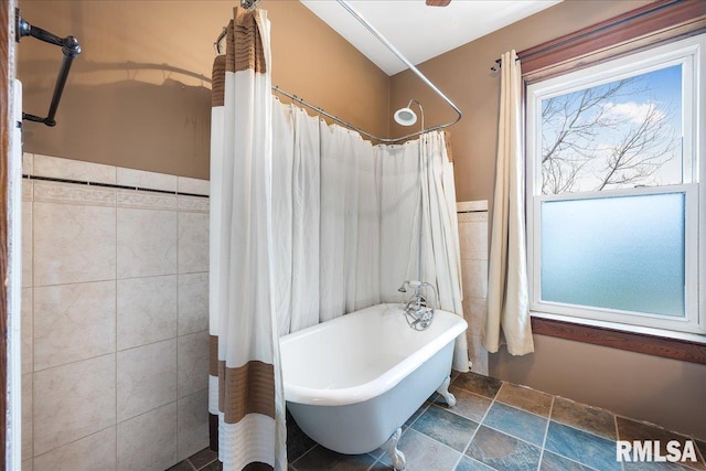 bathroom featuring a freestanding tub, curtained shower, tile walls, stone finish floor, and a wainscoted wall