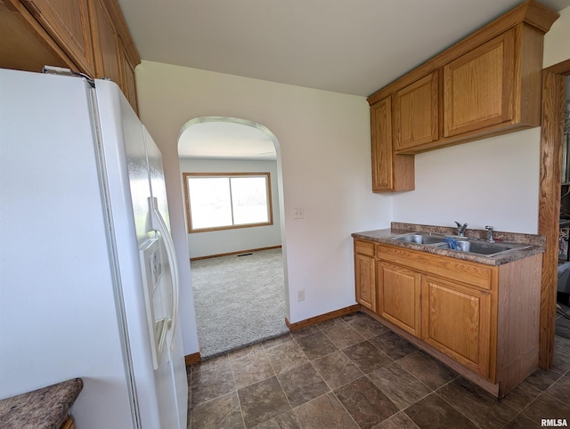 kitchen with dark countertops, a sink, brown cabinets, white fridge with ice dispenser, and arched walkways