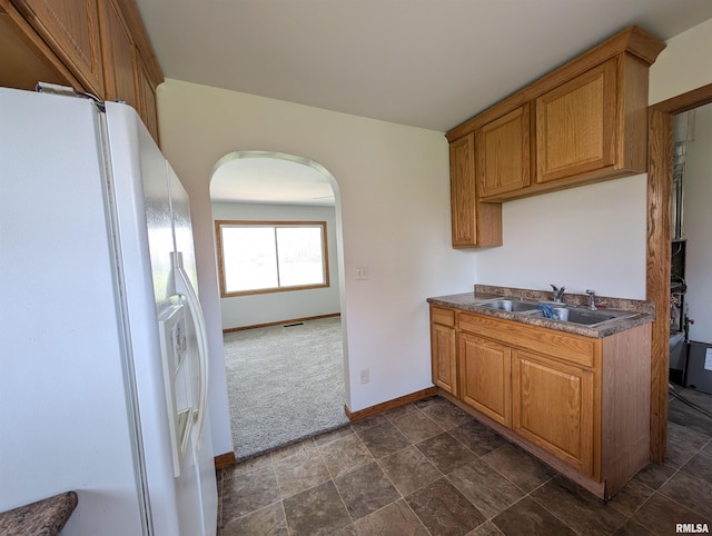 kitchen with arched walkways, a sink, dark countertops, brown cabinets, and white fridge with ice dispenser