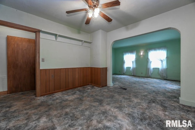carpeted spare room featuring wainscoting, arched walkways, a ceiling fan, and wood walls