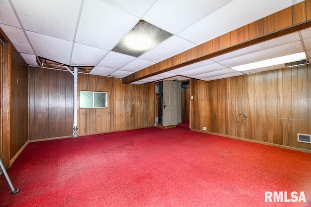 finished below grade area featuring a paneled ceiling, carpet flooring, visible vents, wood walls, and baseboards