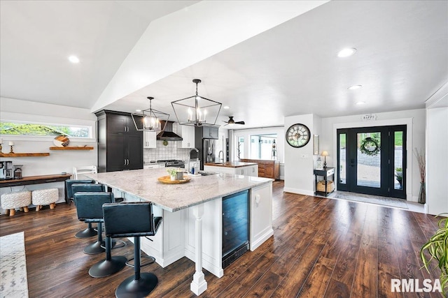 kitchen featuring wall chimney range hood, backsplash, a spacious island, light stone counters, and wine cooler