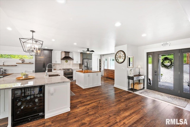 kitchen with beverage cooler, an island with sink, wall chimney range hood, a sink, and appliances with stainless steel finishes