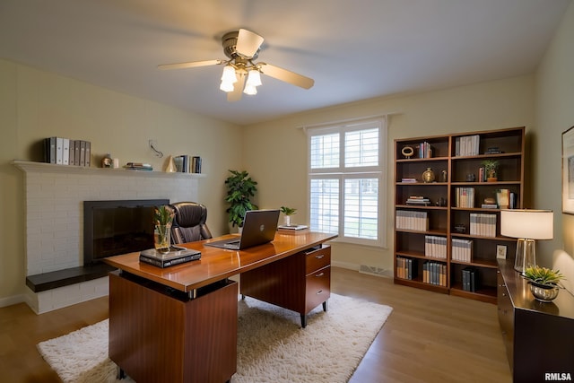 office area with a brick fireplace, a ceiling fan, light wood-style flooring, and baseboards