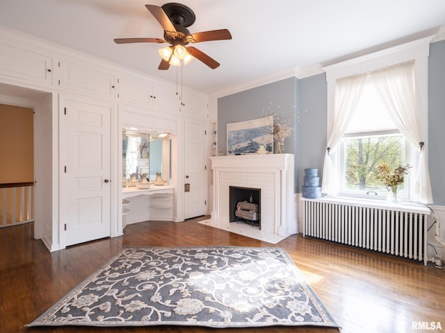 living area featuring radiator, a ceiling fan, a brick fireplace, and wood finished floors
