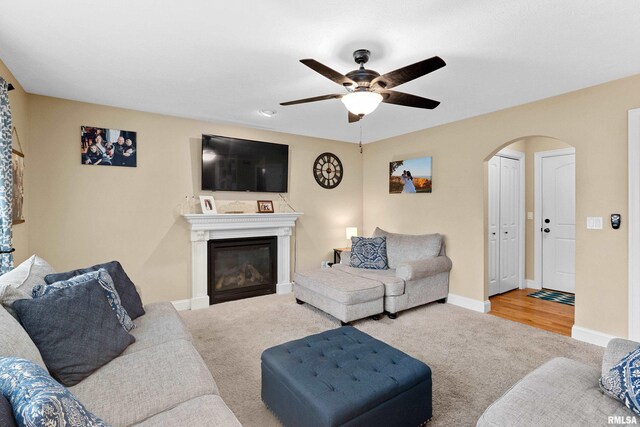 carpeted living area featuring a glass covered fireplace, arched walkways, ceiling fan, and baseboards