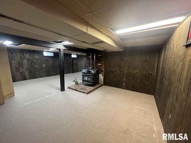 basement featuring visible vents, a wood stove, wooden walls, a paneled ceiling, and carpet