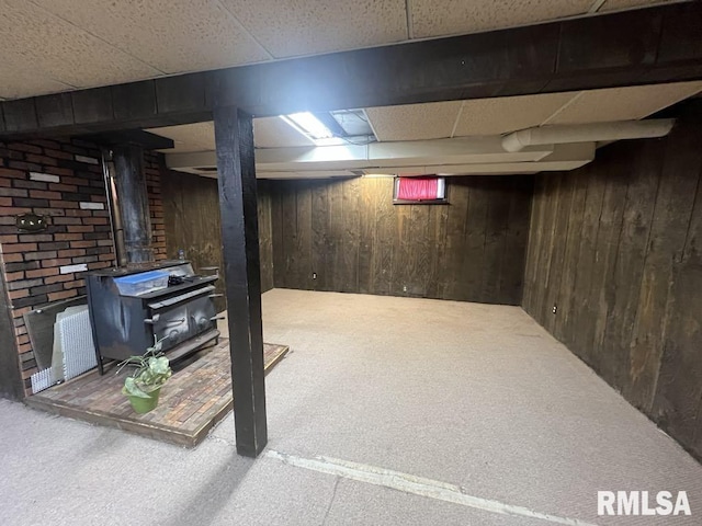 finished below grade area featuring wooden walls, a wood stove, carpet flooring, and a paneled ceiling