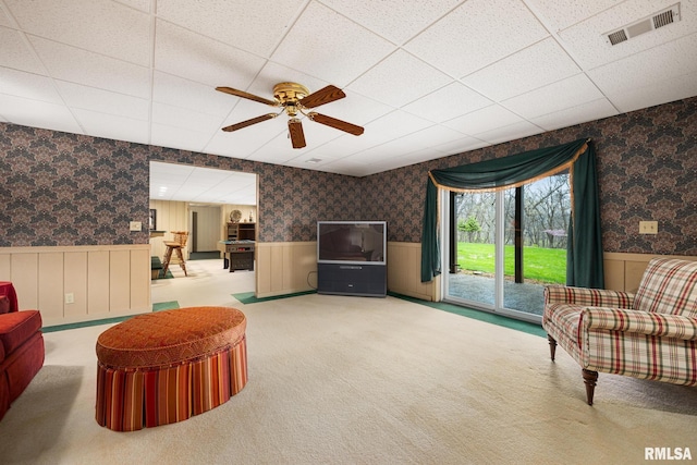 living area with a wainscoted wall, visible vents, a ceiling fan, a drop ceiling, and wallpapered walls