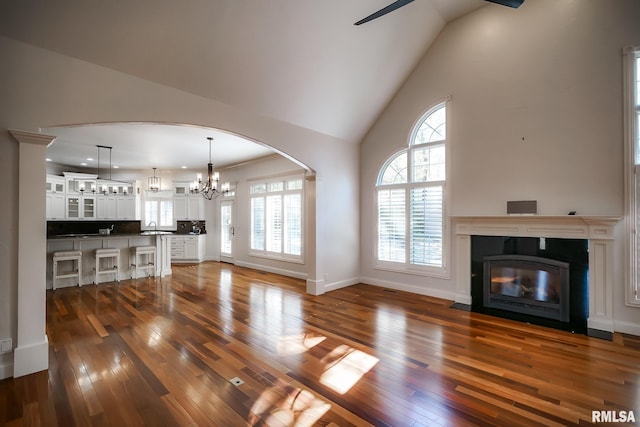 unfurnished living room with ceiling fan with notable chandelier, high vaulted ceiling, dark wood finished floors, arched walkways, and a fireplace with flush hearth