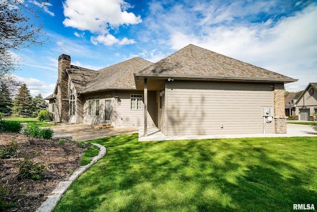 back of house with a chimney, a patio area, a shingled roof, and a lawn