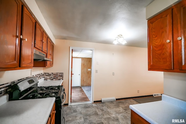 kitchen with stainless steel range with gas cooktop, under cabinet range hood, light countertops, brown cabinetry, and backsplash