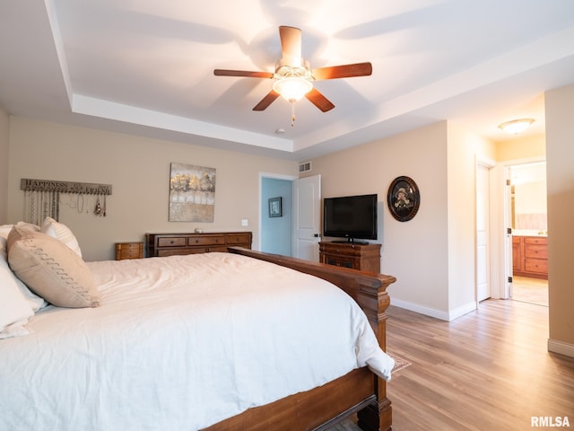 bedroom featuring a raised ceiling, light wood-type flooring, baseboards, ceiling fan, and connected bathroom
