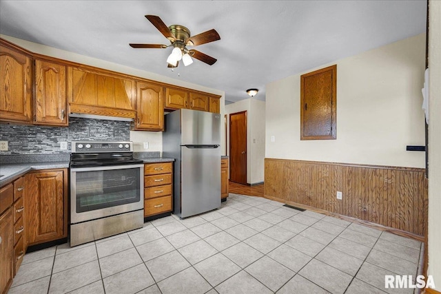 kitchen with appliances with stainless steel finishes, custom exhaust hood, brown cabinetry, ceiling fan, and a wainscoted wall