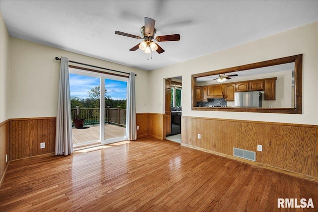 unfurnished living room with wainscoting, light wood-type flooring, a ceiling fan, and wood walls