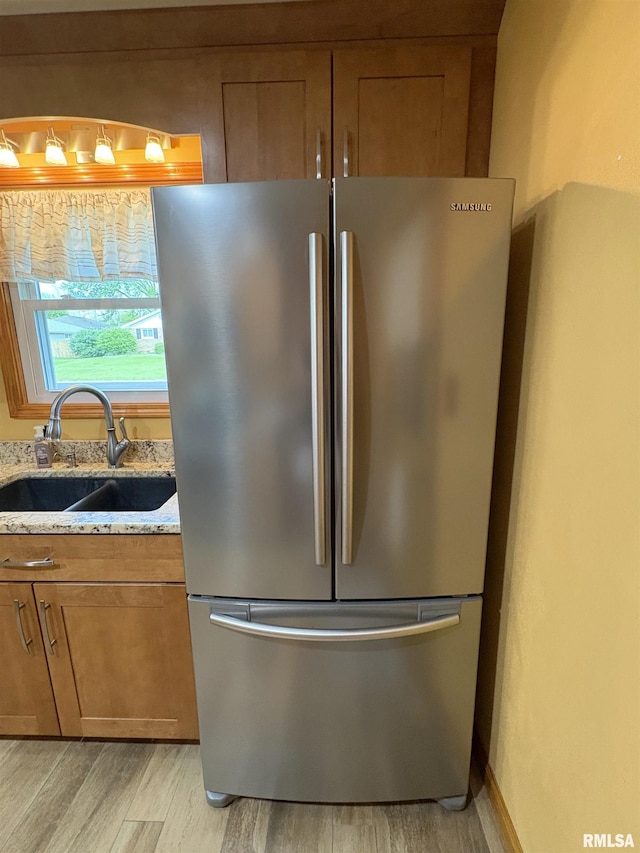 kitchen view of freestanding refrigerator, a sink, brown cabinetry, light wood-style flooring, and light stone countertops