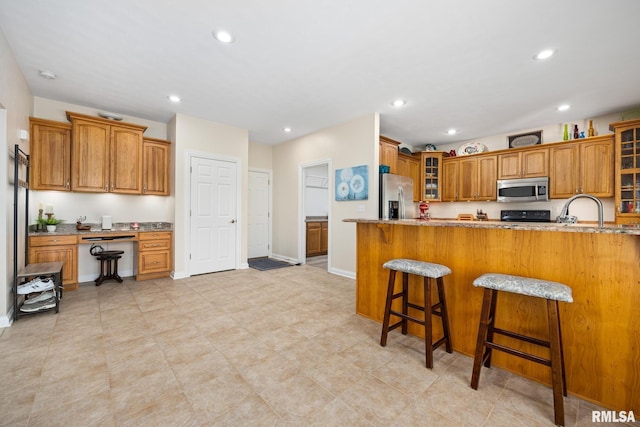 kitchen with appliances with stainless steel finishes, light stone countertops, glass insert cabinets, brown cabinets, and a breakfast bar area