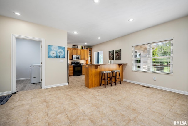 kitchen featuring black range with electric stovetop, stainless steel microwave, a kitchen breakfast bar, brown cabinetry, and recessed lighting