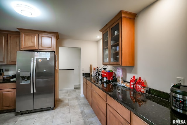 kitchen featuring stainless steel refrigerator with ice dispenser, glass insert cabinets, brown cabinetry, dark stone countertops, and light tile patterned floors