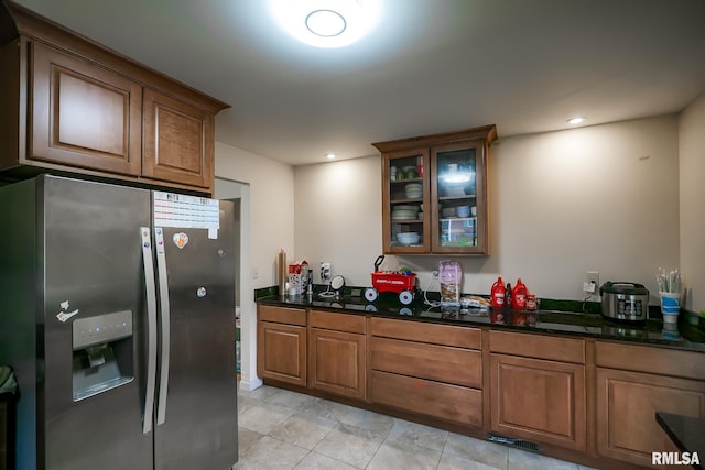 kitchen with stainless steel refrigerator with ice dispenser, glass insert cabinets, brown cabinetry, dark stone countertops, and light tile patterned floors