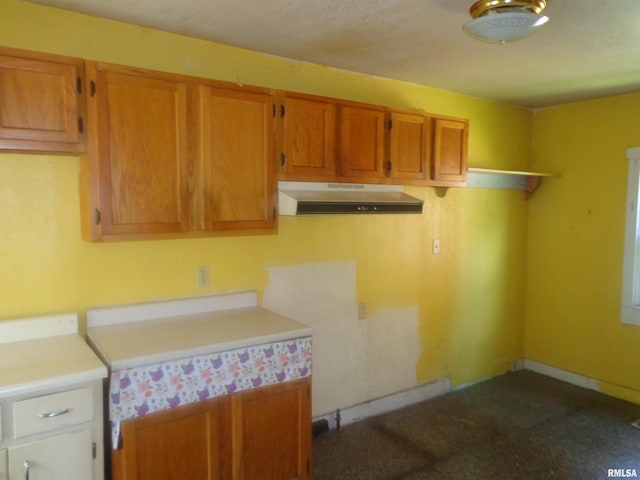 kitchen with baseboards, under cabinet range hood, light countertops, brown cabinets, and dark colored carpet