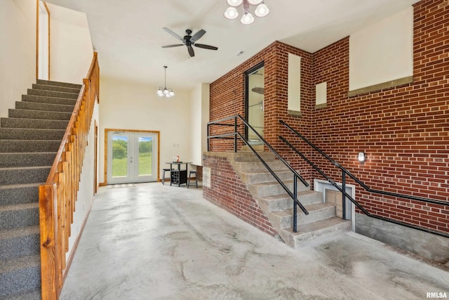 stairs with french doors, concrete flooring, a towering ceiling, brick wall, and ceiling fan with notable chandelier