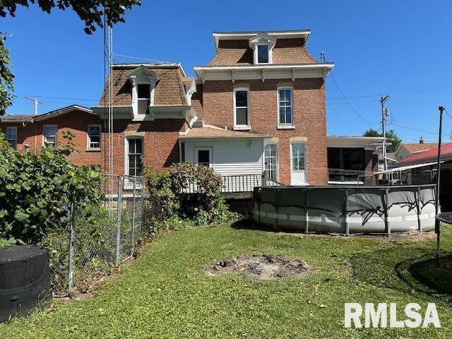 back of house featuring mansard roof, a lawn, roof with shingles, fence, and brick siding