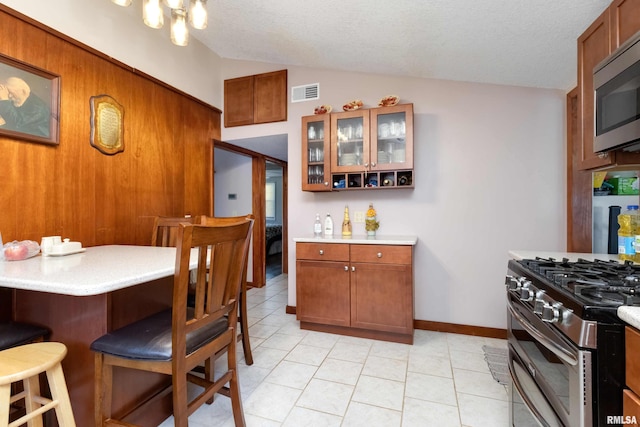 kitchen featuring light tile patterned flooring, a textured ceiling, wooden walls, appliances with stainless steel finishes, and vaulted ceiling