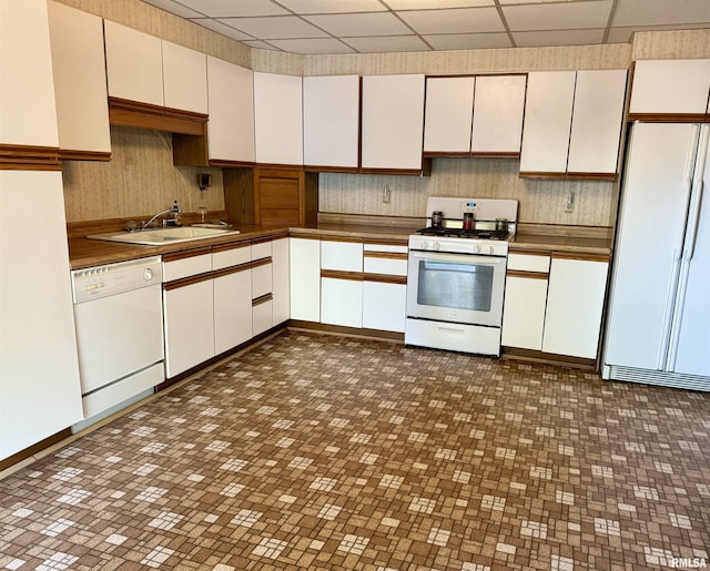 kitchen with dark countertops, white appliances, a paneled ceiling, white cabinets, and a sink