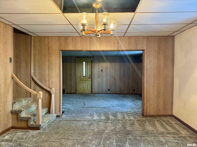 carpeted empty room featuring an inviting chandelier, stairs, a drop ceiling, and wooden walls