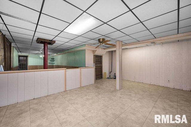 basement featuring a paneled ceiling, ceiling fan, and wooden walls