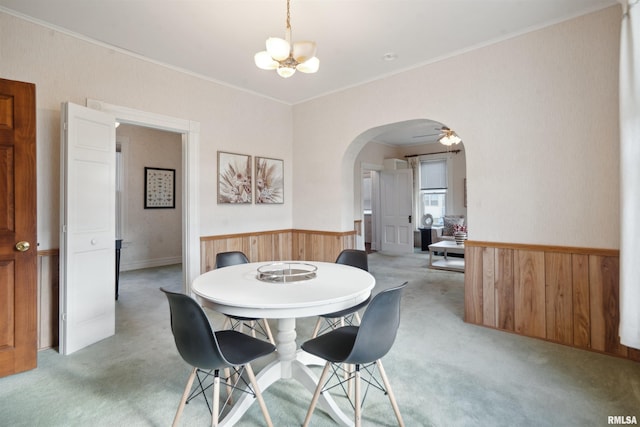 carpeted dining area featuring ceiling fan with notable chandelier, wood walls, and ornamental molding