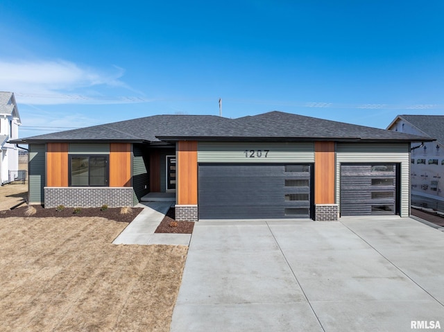 prairie-style home with an attached garage, a shingled roof, concrete driveway, and brick siding