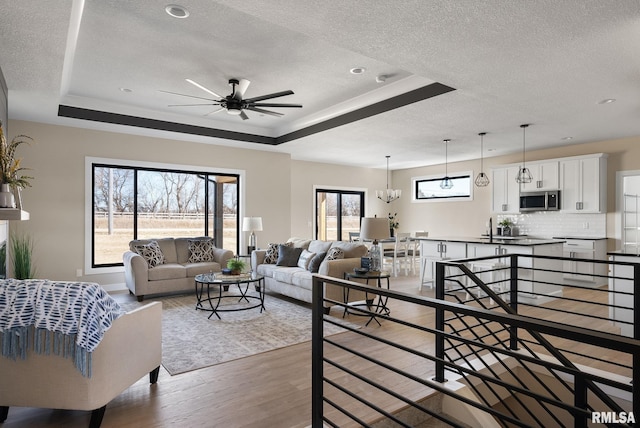 living room with ceiling fan with notable chandelier, a tray ceiling, a textured ceiling, and wood finished floors