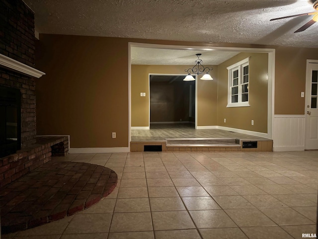 unfurnished living room with a wainscoted wall, ceiling fan, a textured ceiling, tile patterned floors, and a brick fireplace