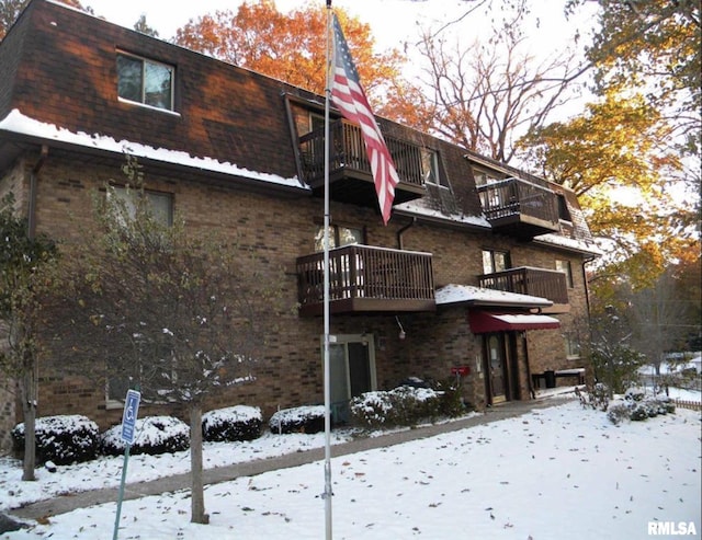 snow covered back of property with a balcony, mansard roof, and brick siding