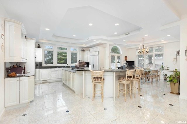 kitchen featuring white cabinets, white built in fridge, a kitchen island with sink, and a tray ceiling