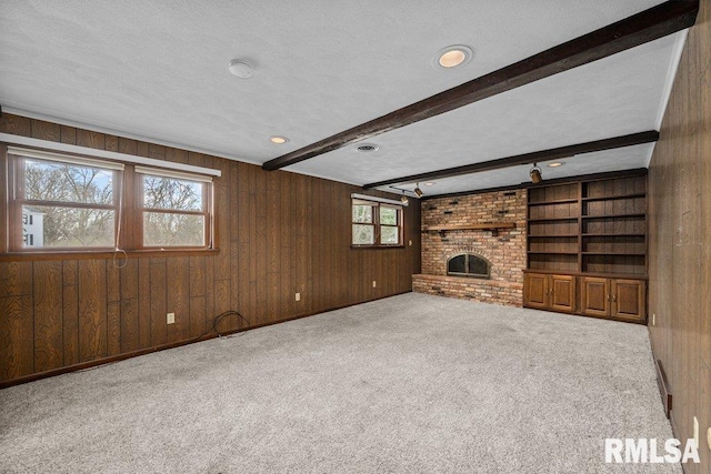 unfurnished living room with wood walls, a brick fireplace, a textured ceiling, beamed ceiling, and light colored carpet