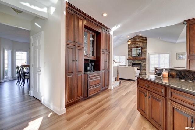 kitchen with dark stone counters, vaulted ceiling, ceiling fan, light hardwood / wood-style flooring, and a stone fireplace