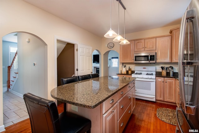 kitchen with appliances with stainless steel finishes, light brown cabinetry, dark stone counters, decorative light fixtures, and a center island