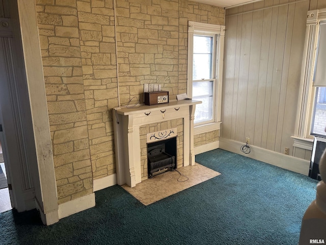 unfurnished living room featuring a stone fireplace, a wealth of natural light, wooden walls, and dark carpet