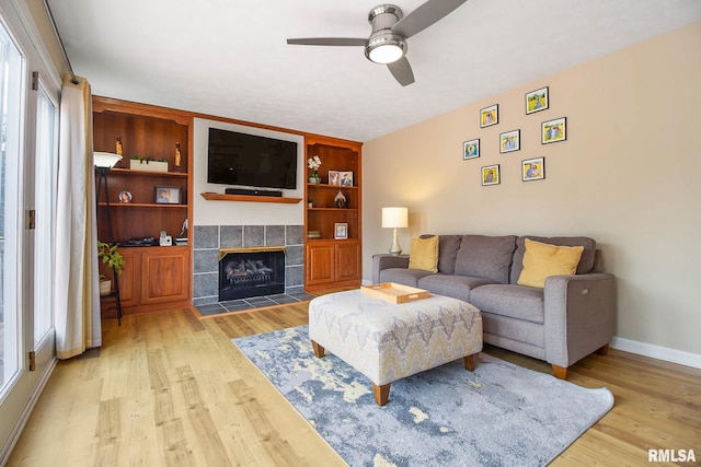 living room with a tiled fireplace, ceiling fan, built in features, and light hardwood / wood-style floors