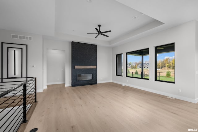 unfurnished living room with a large fireplace, a tray ceiling, light hardwood / wood-style flooring, and ceiling fan