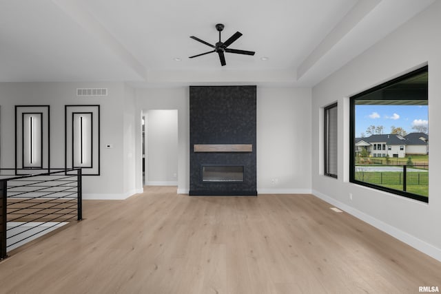 unfurnished living room with ceiling fan, a large fireplace, a tray ceiling, and light hardwood / wood-style flooring