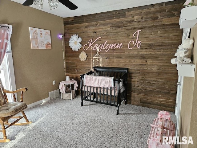 carpeted bedroom featuring a nursery area, wooden walls, and ceiling fan