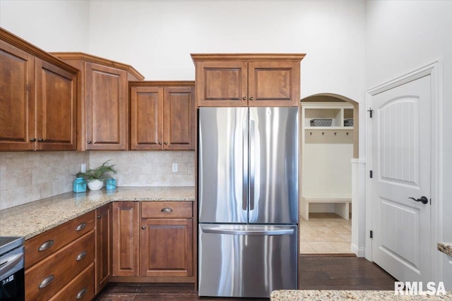 kitchen featuring light stone counters, brown cabinetry, arched walkways, freestanding refrigerator, and tasteful backsplash