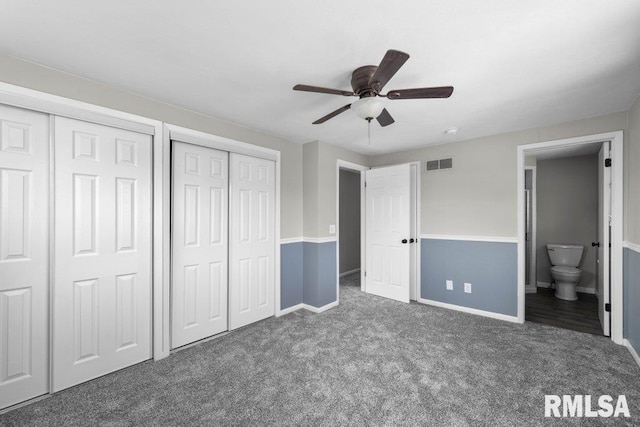 unfurnished bedroom featuring ceiling fan, dark carpet, ensuite bath, and two closets