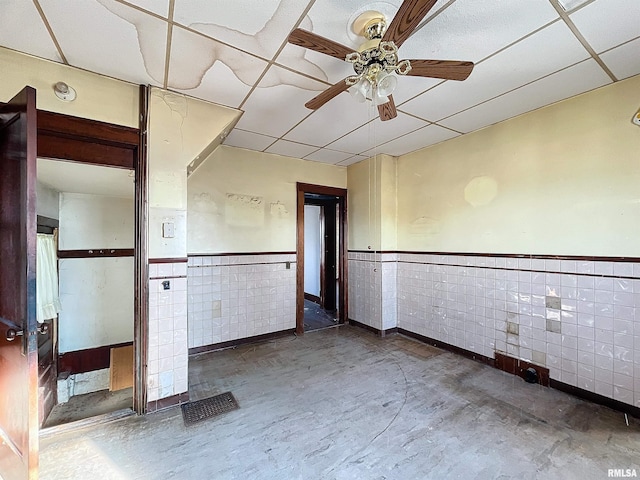 unfurnished room with a paneled ceiling, ceiling fan, tile walls, and concrete floors